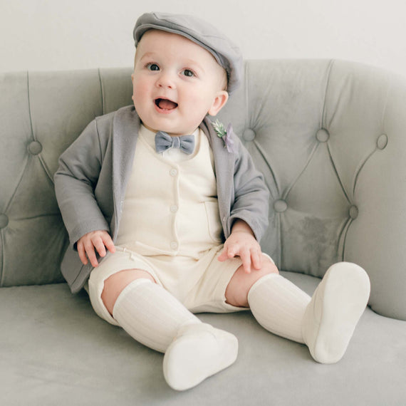 Baby boy sitting on a couch wearing the Ezra 4-Piece Suit with matching Newsboy Cap, heather velvet bow tie and boutonniere