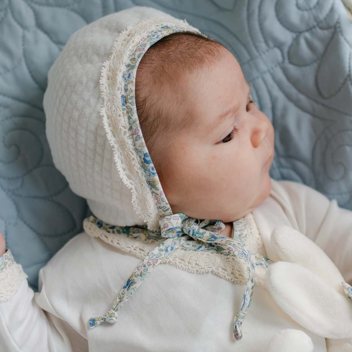 Baby lying in a wicker bassinet on a blue quilt, wearing the Petite Fleur Quilted Bonnet with floral trim and ecru lace, holding a soft bunny toy.