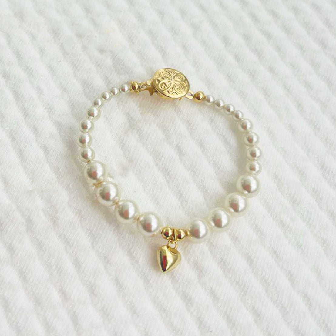 Cream pearl baby bracelet with gold plated heart charm on a white textured background
