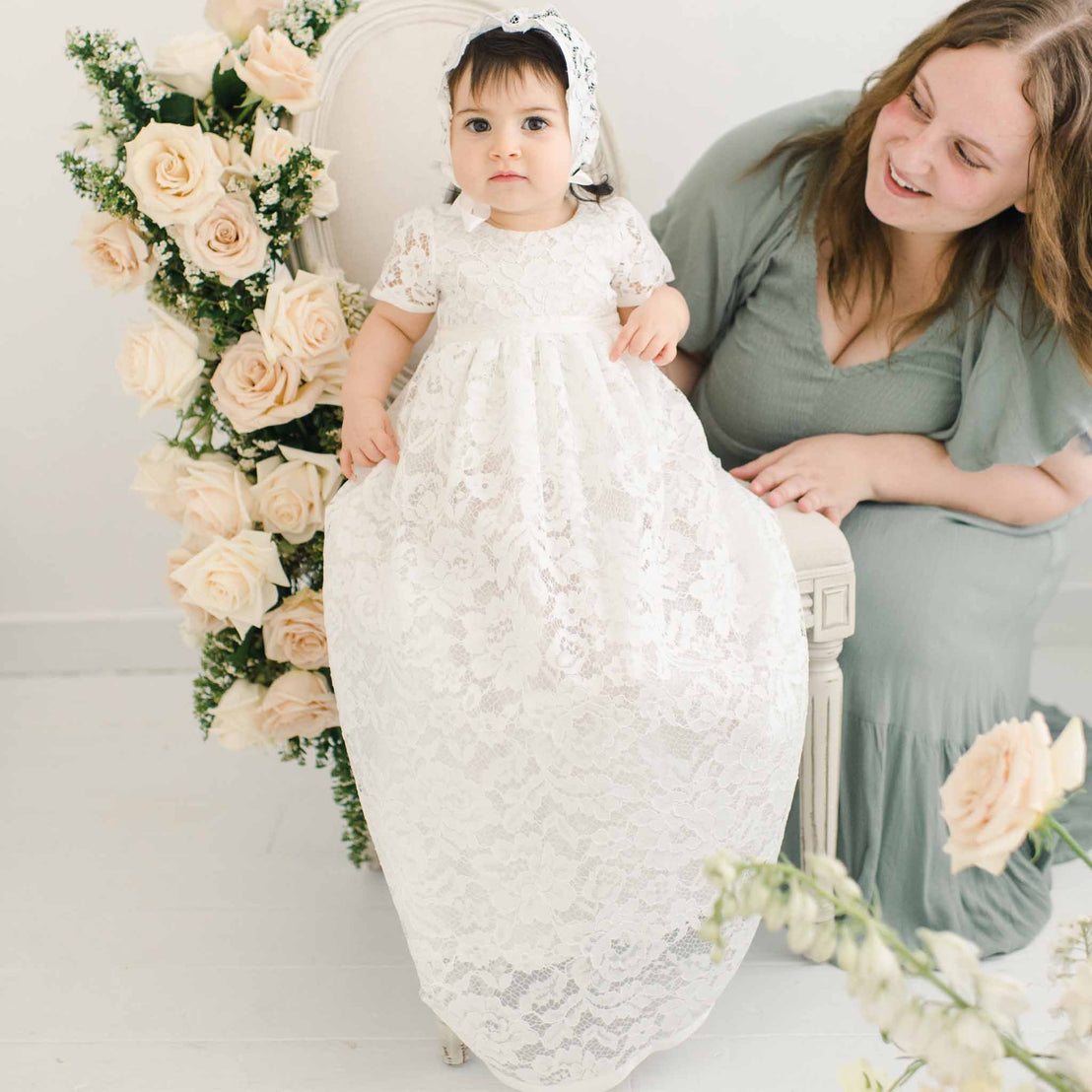 A woman in a gray dress smiles at a baby in a Rose Christening Gown & Bonnet, sitting on her lap. They are surrounded by cream roses in a light, airy room.