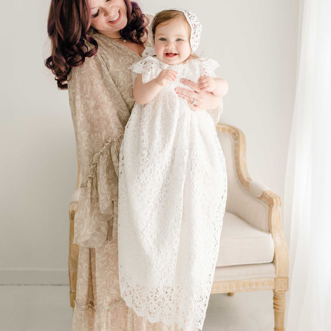 Baby girl with her mother. She is wearing the Lola Christening Gown and Bonnet, a baptismal gown made with a cotton undergown in light ivory featuring an all-over lace overlay.