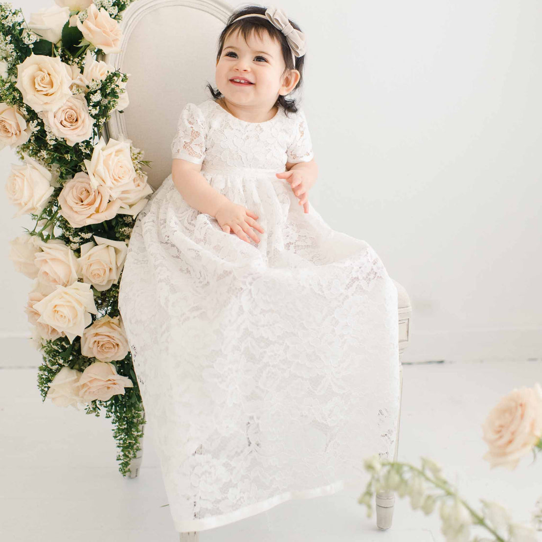 A toddler girl with a joyful smile, wearing an ivory lace Rose Christening gown and bonnet, sits on an elegant chair adorned with pink roses in a light, bright room.