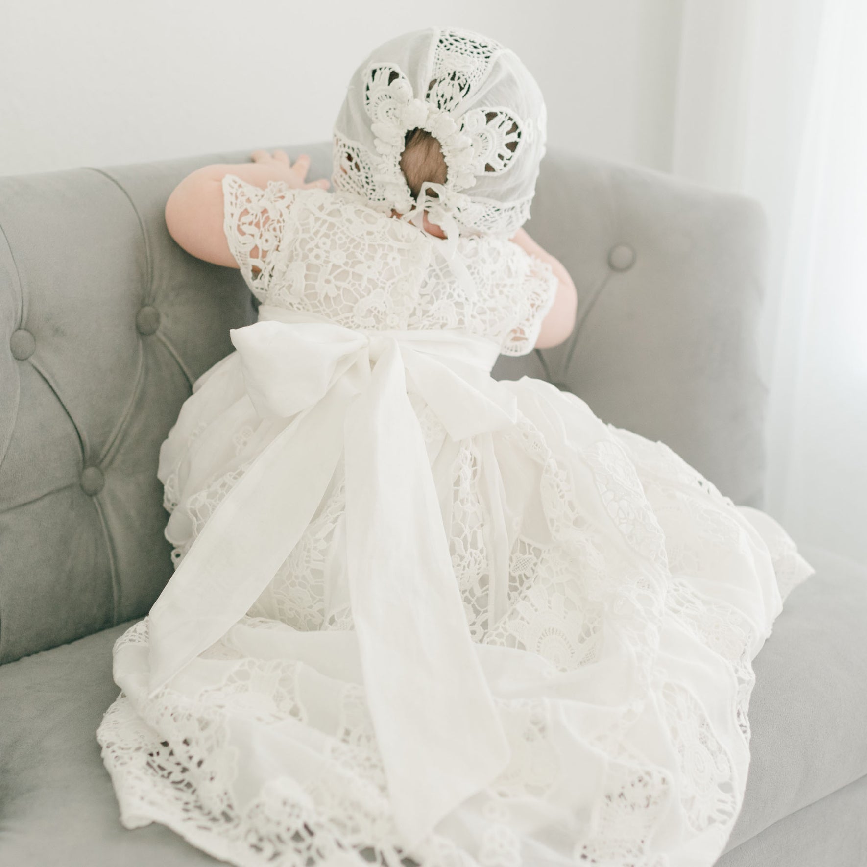 A baby in the Grace Christening Gown & Bonnet, which features an intricate lace design and a large silk sash at the back, leans against a light gray tufted sofa. The baby is facing away, highlighting the detailed lace pattern of the outfit.