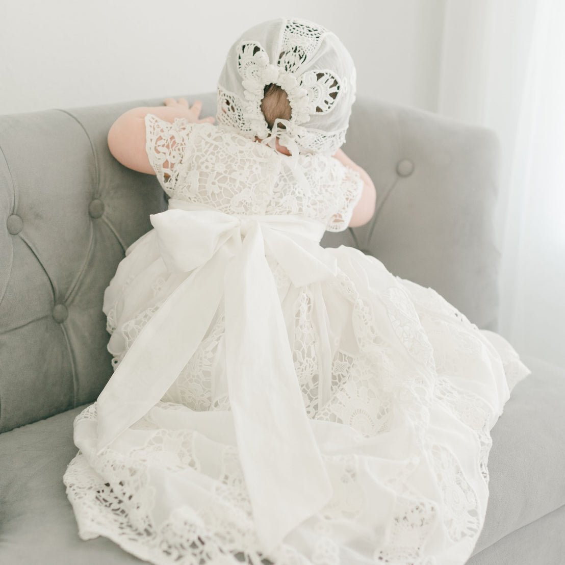 A baby, dressed in a white lace outfit with a large bow at the back and wearing the Grace Lace Bonnet—an ivory baby bonnet partially decorated with flowers—is kneeling on a grey tufted sofa, facing away from the camera. The background is softly lit.