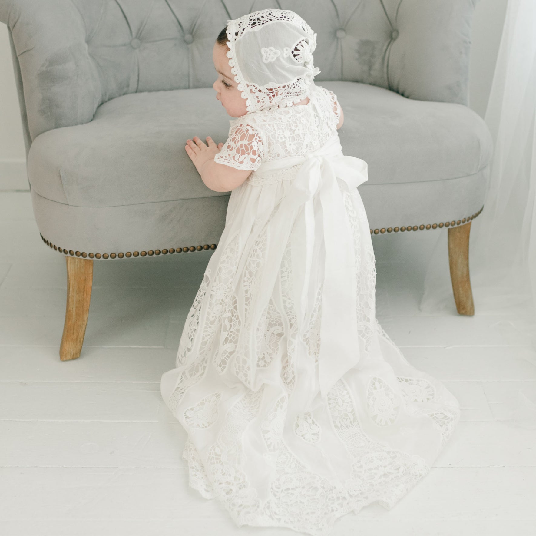 A young child stands facing a gray upholstered chair, dressed in the exquisite Grace Christening Gown & Bonnet. The detailed lace gown features a long train and is complemented beautifully by a matching bonnet adorned with delicate patterns. A silk sash adds an elegant touch to the outfit. The scene unfolds against a backdrop of a white floor and sheer curtains.