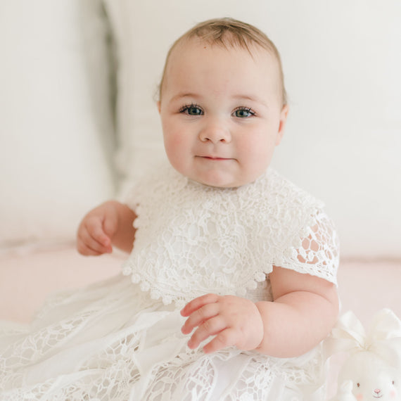 A baby wearing a Grace Lace Christening Bib sits upright, showcasing the intricate lace detailing. The baby has light skin and a slightly smiling expression. In the background, neutral-colored pillows create a soft ambiance, and a small white plush toy is partially visible at the baby's side.