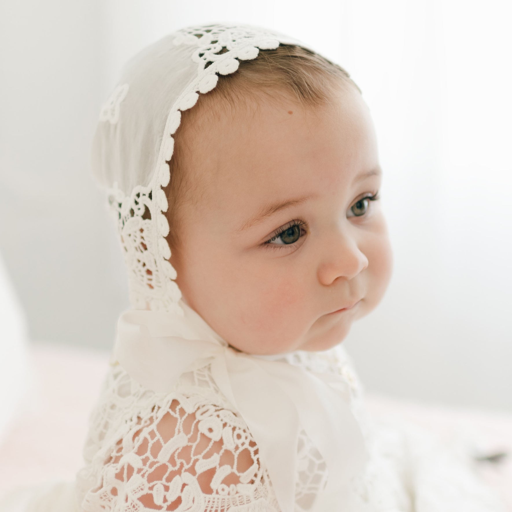 A baby wearing the Grace Lace Bonnet and a matching ivory outfit gazes off to the side. The bonnet features intricate details and scalloped edges. The baby's cheeks are slightly flushed, and natural light softly illuminates the scene.