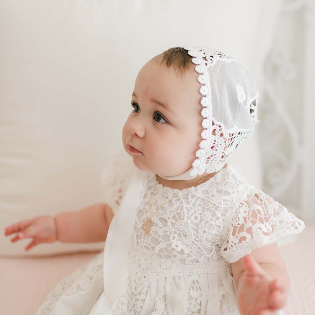 A baby wearing a Grace Lace Bonnet and a detailed white lace dress sits against a light-colored backdrop. The baby faces slightly to the left with hands raised, softly looking upward. The dress is adorned with intricate lace patterns.