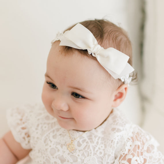 A baby with a delicate Grace Christening Headband adorned with a white bow, dressed in a beautiful white lace outfit. The baby has short hair and a faint smile, looking slightly to the side. The background is light and out of focus, adding to the serene charm of the moment.