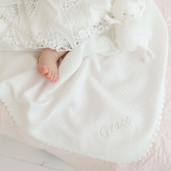 A baby rests on a handmade Grace Personalized Blanket, which is beautifully embroidered with the name "Grace." The soft cotton lace cloth partially covers the baby's tiny feet, and a small white stuffed animal lies next to the baby on the blanket, forming an exquisite personalized heirloom piece.