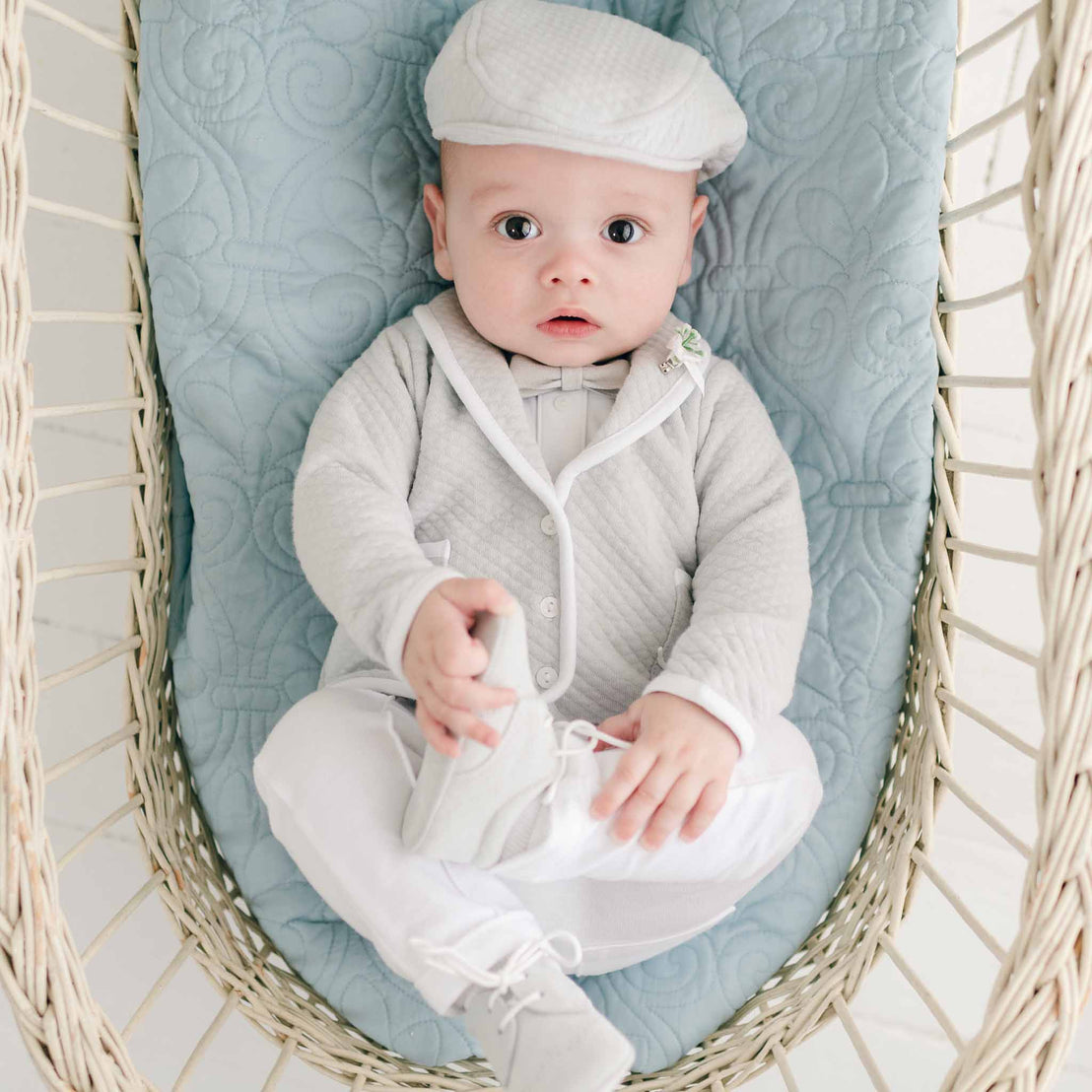 A baby, dressed in the Grayson 3-Piece Suit, crafted with care in the USA, is nestled charmingly in a basket lined with blue fabric. With an endearing gaze towards the camera, the little one holds one of his shoes in a tiny hand.