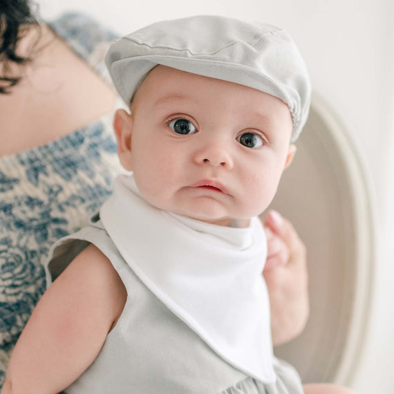 A baby wearing the Grayson Linen Romper, matching Newsboy Cap, and Grayson Bib Bandana Bib, made from white French Terry cotton, is sitting on the lap of a person with dark hair. The baby has large eyes and is looking directly at the camera. The person holding the baby is partially visible and dressed in a patterned top.