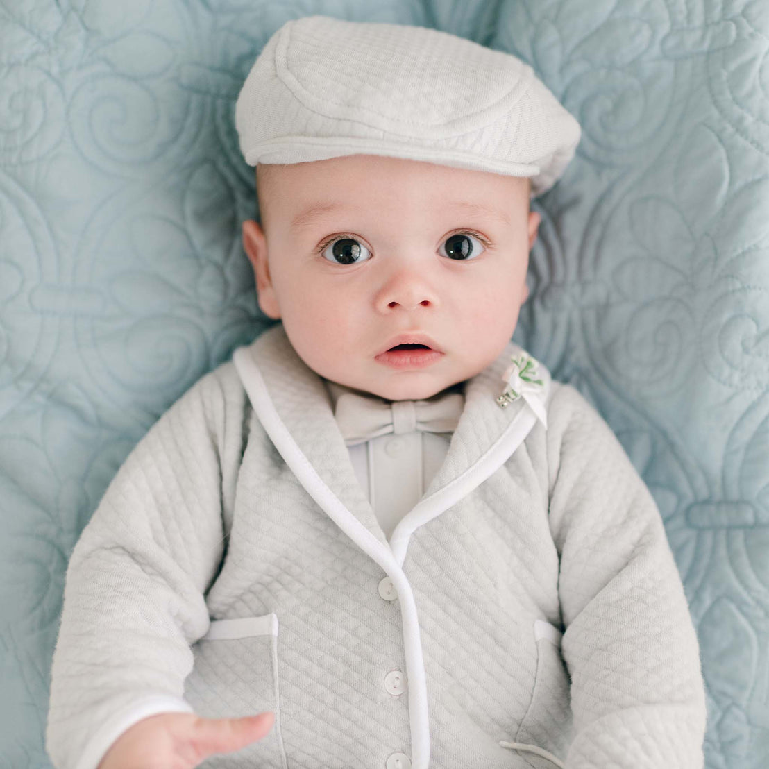 A baby, wearing the delightful Grayson 3-Piece Suit with a textured light gray outfit and matching cap, relaxes on a quilted soft blue background. His wide-open eyes exude curiosity, adorned with a bow tie and small boutonniere for an elegant touch.