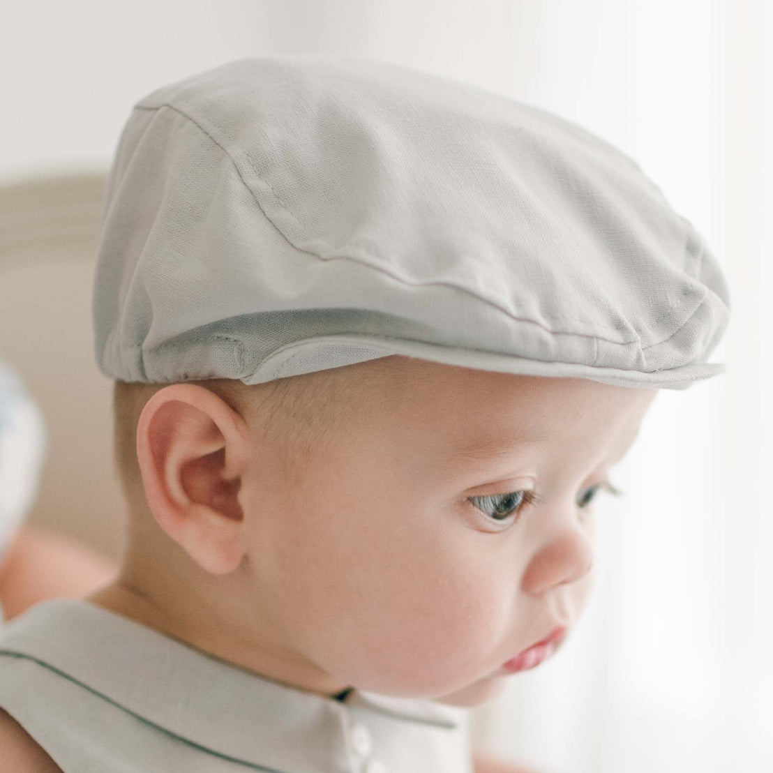 A baby wearing a Grayson Linen Newsboy Cap and the Grayson Linen Romper looks to the right. The background is softly blurred.