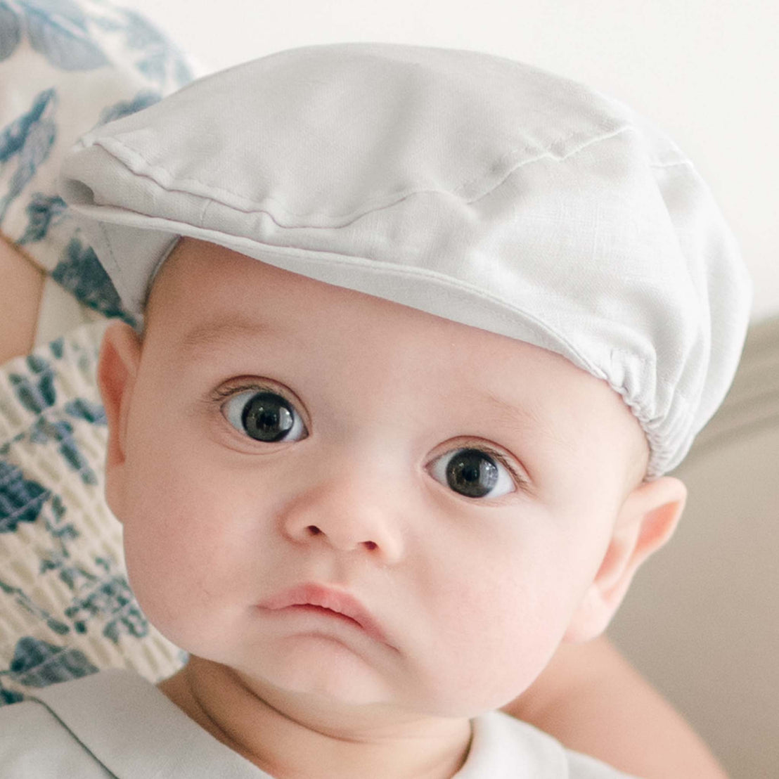 A baby wearing a Grayson Linen Newsboy Cap is looking at the camera. The baby has large eyes and rosy cheeks. Part of an adult's arm and patterned clothing are visible in the background.