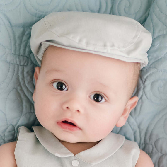 A baby wearing a Grayson Linen Newsboy Cap and matching Grayson Linen Romper lies on a light blue quilted surface. The baby has large, dark eyes and slightly parted lips, looking directly at the camera.