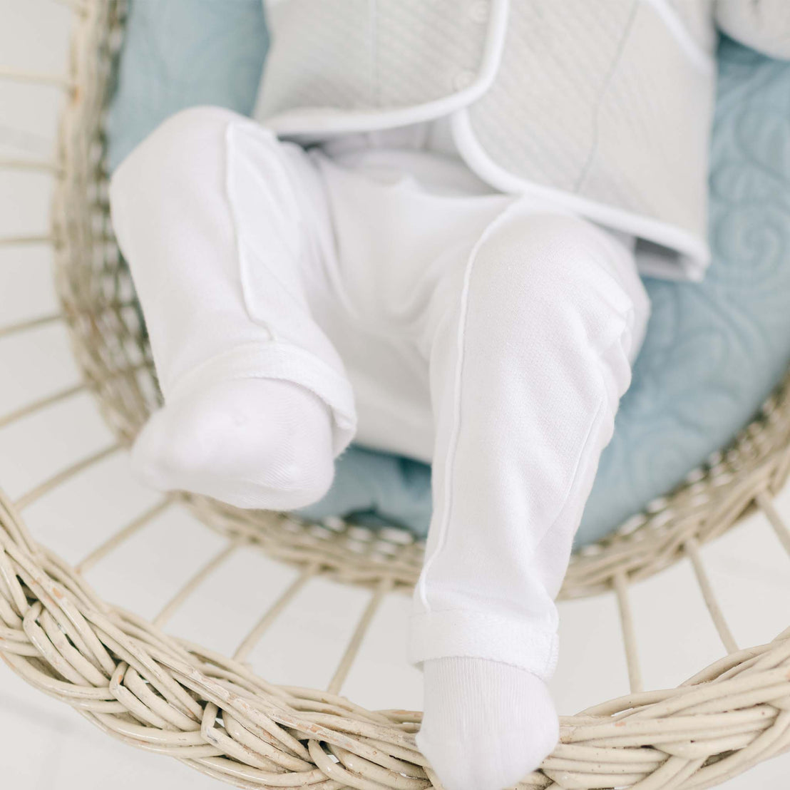 A baby dressed in white pants and a gray shirt, reminiscent of a comfy, laid-back alternative to the Grayson 3-Piece Suit, is reclining in a wicker bassinet with a light blue cushion. The baby's face is not shown in the image.