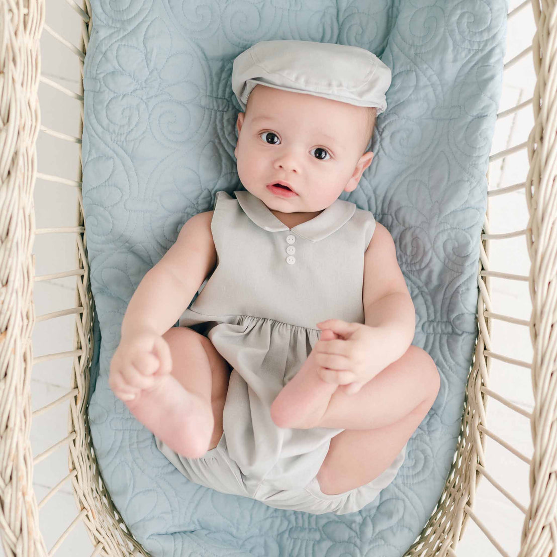 A baby lies in a wicker bassinet on a light blue quilted blanket, wearing the charming Grayson Linen Romper and cap. The little one looks up with wide eyes, one hand holding a foot, exuding the cozy appeal of handmade baby clothes.