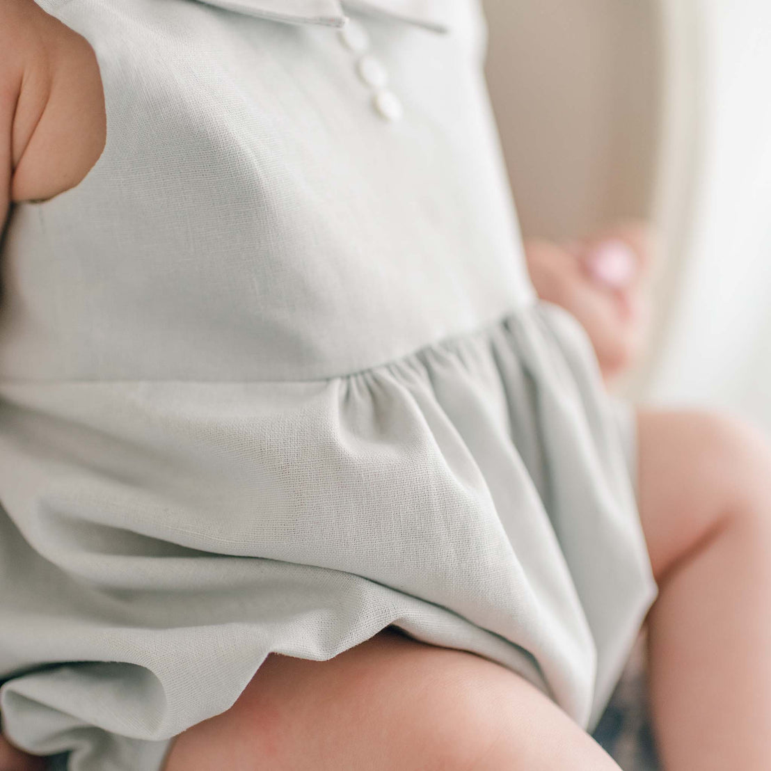 A baby wearing a Grayson Linen Romper and matching Newsboy Cap lies in a wicker bassinet with a quilted blue blanket. The baby looks up at the camera with wide eyes and has both hands reaching toward their feet.