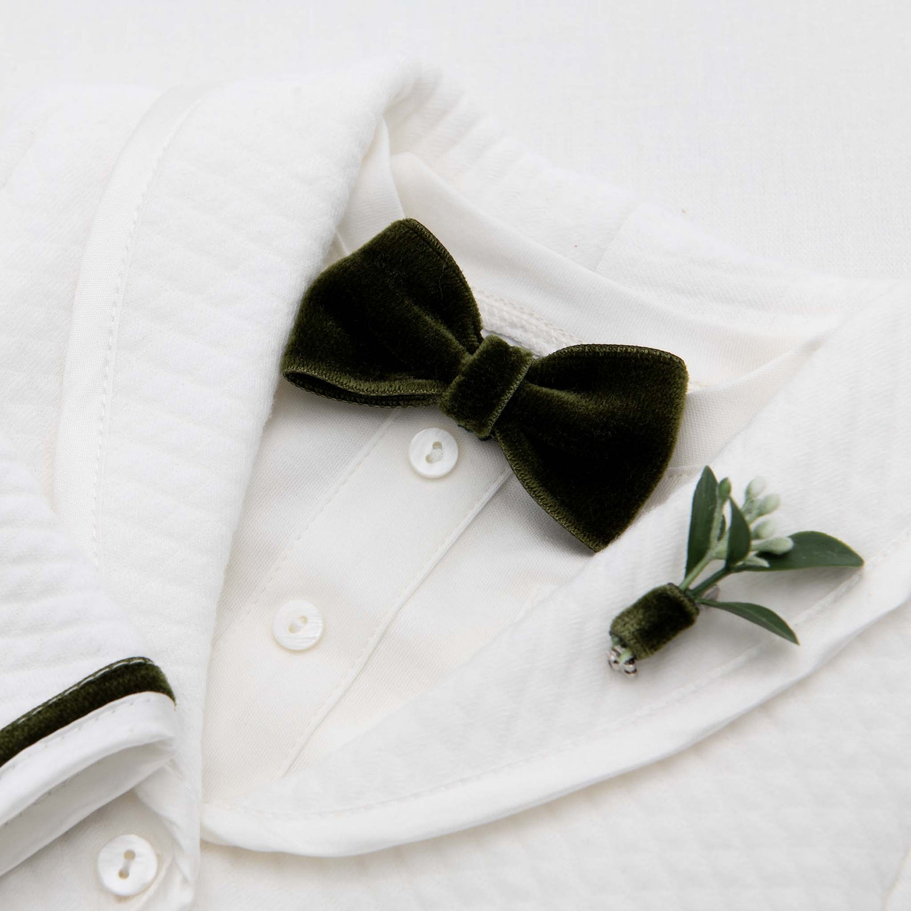 The Noah Green Bow Tie & Boutonniere set showcases a luxurious dark green velvet bow tie, handmade in the USA, elegantly paired with a small boutonniere of greenery and a white flower bud, pinned on the left lapel of a textured white dress shirt. It's the perfect holiday accessory for any elegant occasion.