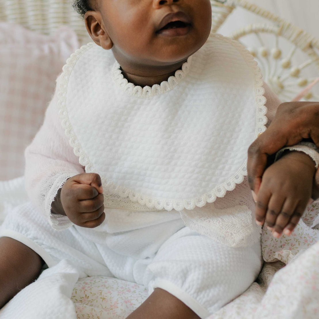 Baby wearing the Hailey Bib and matching romper, sitting on a patterned blanket. Details show quilted cotton adorned with an ivory edge lace. Perfect baptism gift from godparents.