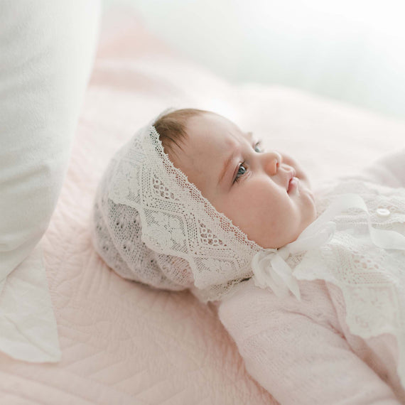 A baby wearing a Hailey Knit Bonnet and an outfit with lace details is lying on a pink knit acrylic quilted blanket, gazing to the side. A white pillow is partially visible on the left side of the image.