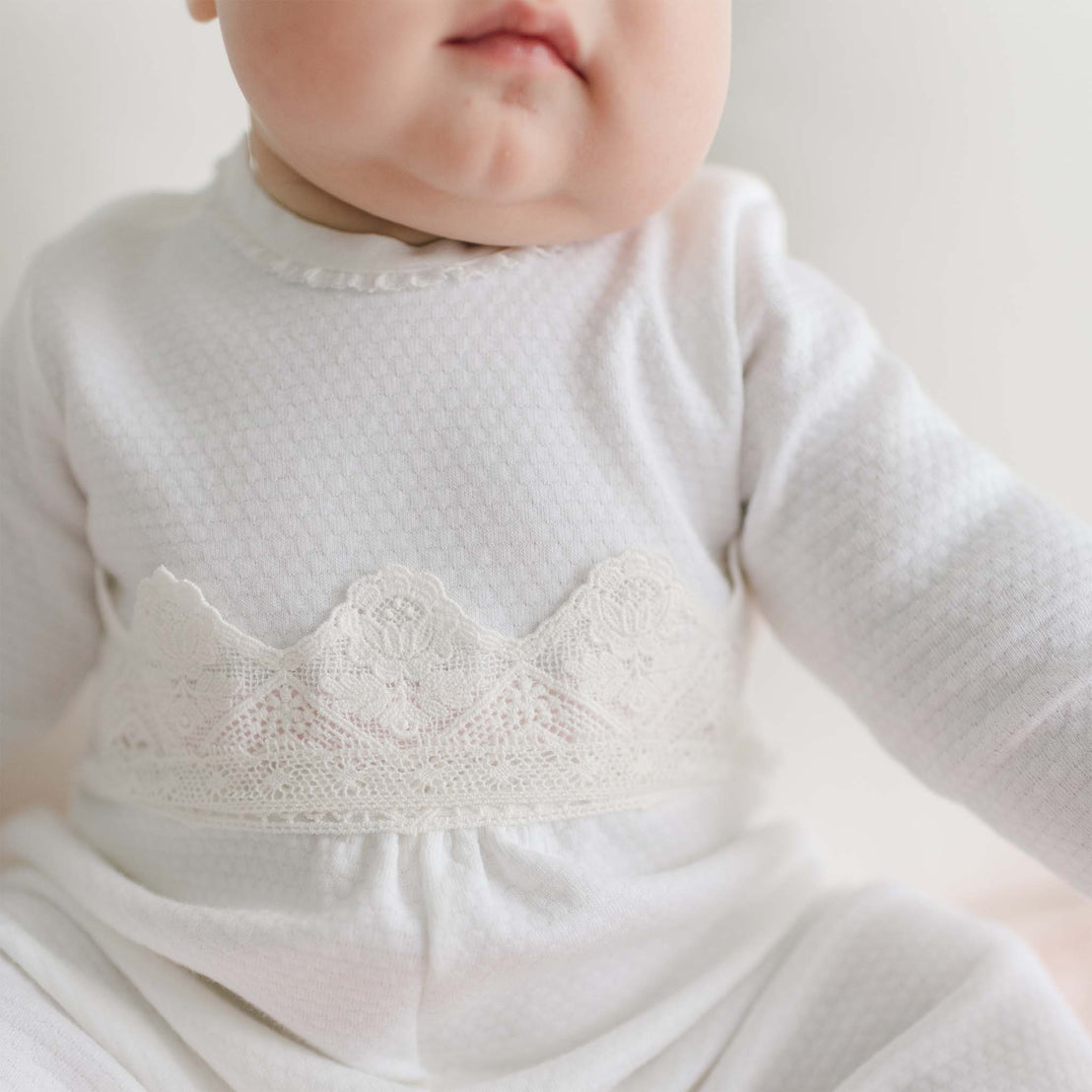 A close-up image of a baby dressed in a textured white 100% cotton Hailey Romper with long sleeves. The outfit features a delicate lace pattern across the chest. Only the lower part of the baby's face and upper body are visible against a white background, showcasing this heirloom baby attire.