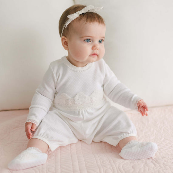 A baby is sitting on a light pink quilted surface, wearing a Hailey Romper—a white outfit with lace details and a matching headband with a bow. The baby's eyes are looking slightly to the side. The background is plain white, highlighting the timeless elegance of the ensemble.