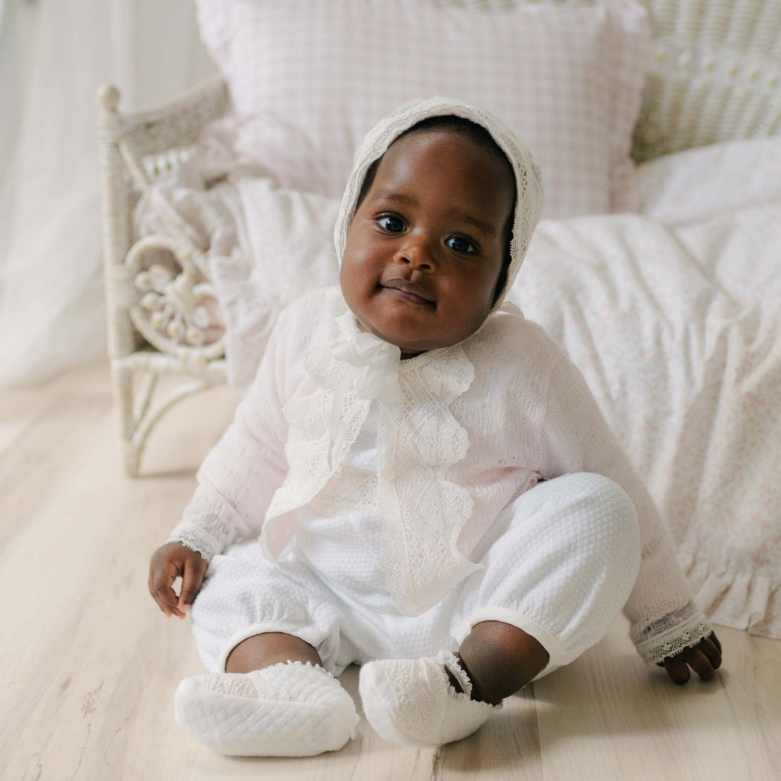Baby in the Hailey Knit Sweater sitting on a light wooden floor with a white wicker chair in the background. Details of the sweater show the pink knit with matching light ivory laces. Perfect for unique baby gifts. 