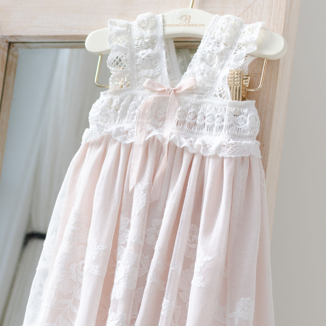 An elegant pink Charlotte Christening Gown & Bonnet for a little girl, featuring delicate cotton lace detailing and a satin ribbon at the chest, hanging on a wooden hanger against a soft background.
