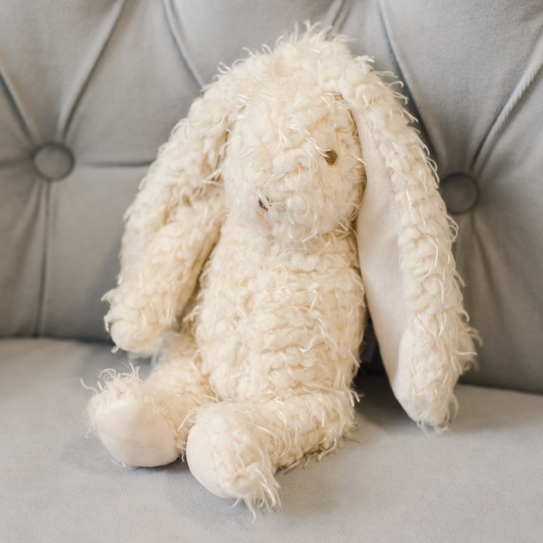 The Grace Bunny, with its cream-colored textured fur-like appearance and irresistibly soft, fluffy fur, sits gracefully on a light gray cushioned surface. Its long ears make it an ideal baby gift that invites endless cuddles.