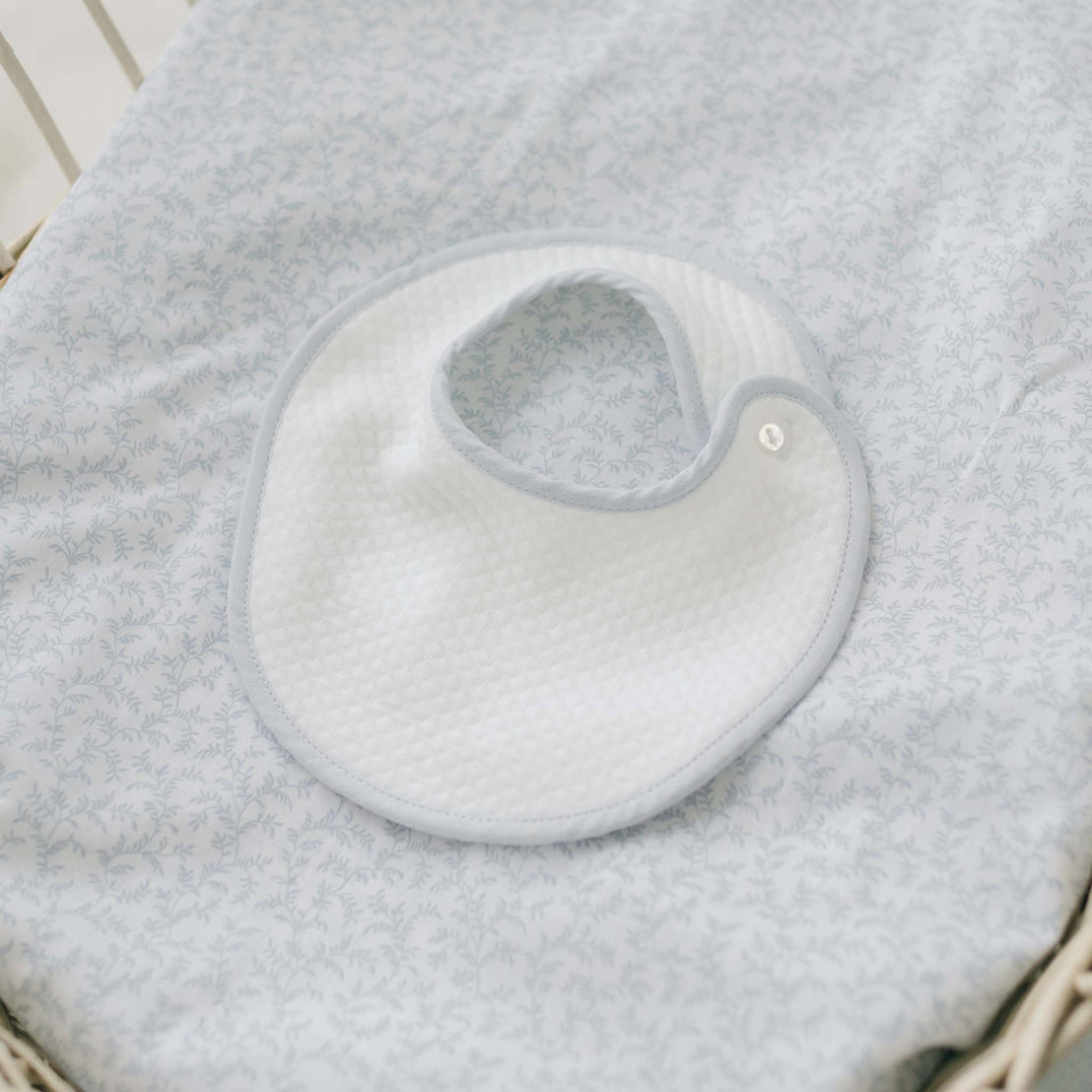 A white Harrison Christening Bib, made from textured cotton and adorned with light blue trim, rests on a light blue patterned fabric. This handmade bib showcases a rounded shape and is secured with a snap fastener at one end.