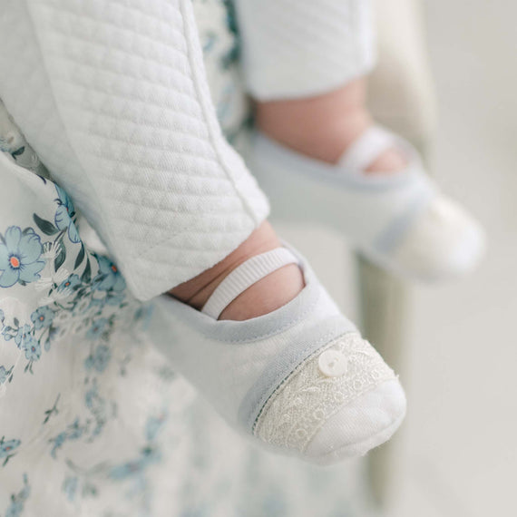 Close-up of a baby's legs and feet, wearing white quilted pants and light blue Harrison Christening Booties with lace details and a small button on each. The baby appears to be sitting on a floral-patterned fabric.