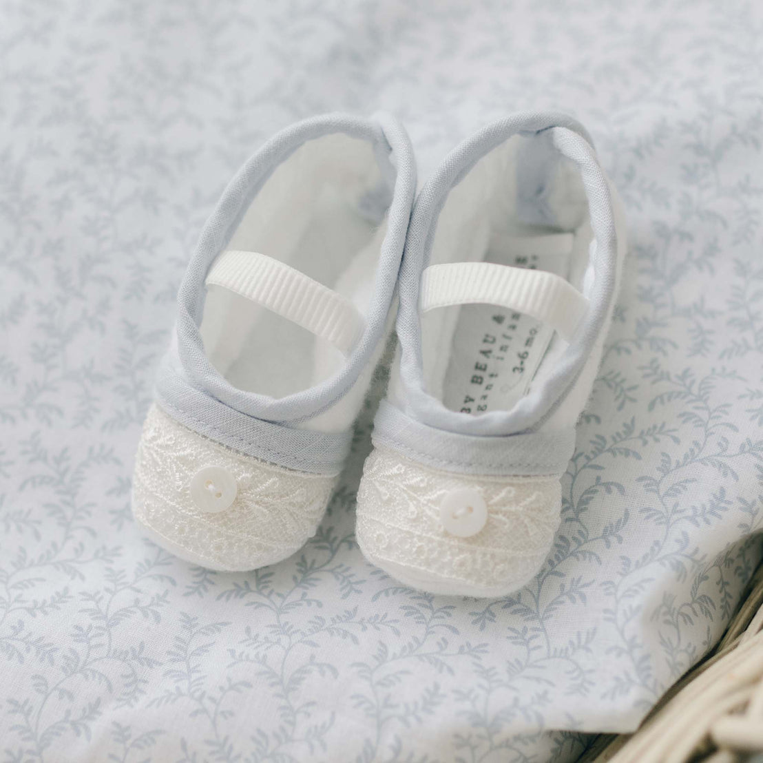 The image showcases a pair of Harrison Christening Booties, featuring white embroidered toes and light blue fabric. These baby shoes, ideal for a baptism outfit, are arranged on a subtly patterned floral fabric and secured with a white strap.