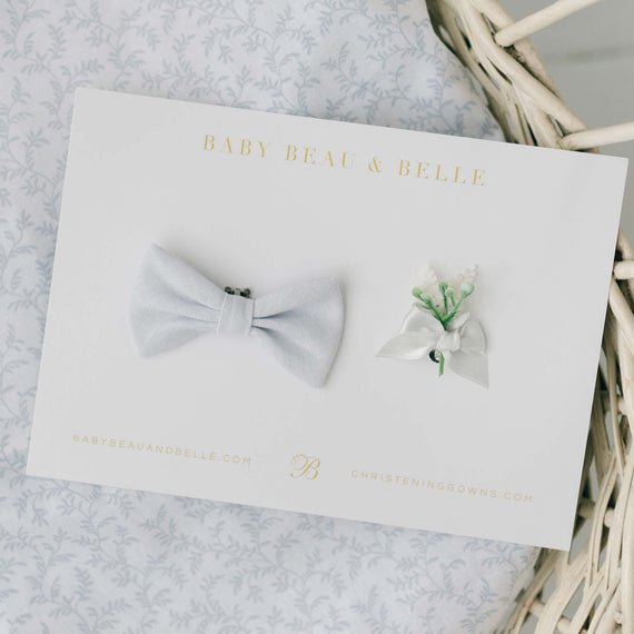 A display card from "Baby Beau & Belle" showcases the Harrison Linen Bow Tie & Boutonniere, featuring a charming light blue linen bow tie and a small white floral boutonniere, both handmade in the USA. The card also includes the brand's website and details about christening gowns, with the items elegantly placed on a light-patterned background.
