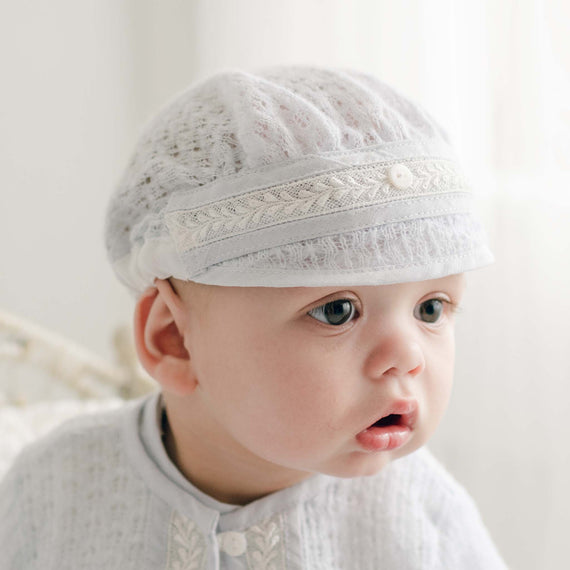 A baby is wearing a white lace cap and white clothing with an embroidered pattern. Sporting a curious expression, the baby glances slightly to the right. The softly lit background provides a gentle, neutral setting, perfectly complementing the handmade USA quality of the Harrison Blue Knit Christening Hat.
