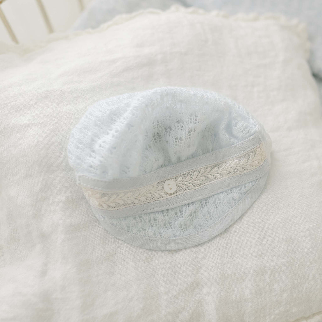 The Harrison Blue Knit Christening Hat, featuring a delicate, light blue knit with a decorative white band and a small white button, lies on a white, textured fabric surface. Its soft and intricate design suggests it is intended for an infant.