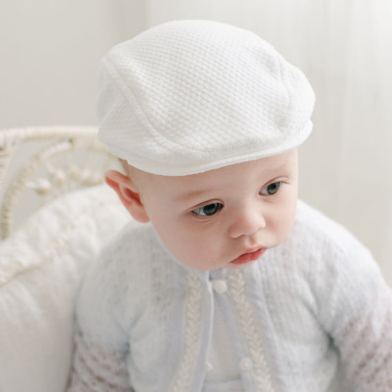 A baby wearing a white knitted outfit and the Harrison Textured Newsboy Cap sits on a light-colored surface. The baby is looking slightly to the side with a neutral expression. The background is soft and blurred.