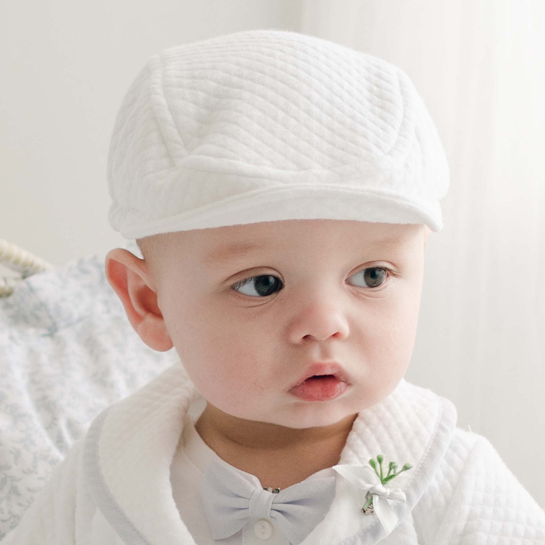 A baby donning the Harrison Newsboy Cap in quilted cotton, along with a matching white outfit and bow tie, is gazing slightly to the side. The softly blurred background enhances the calm and serene atmosphere.