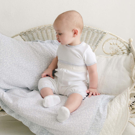 A baby wearing the Harrison Short Sleeve Christening Romper sits on a wicker cushioned seat set against a light-colored background. The baby boy, clad in socks, rests one hand on the cushion while his feet rest on a blanket. This adorable scene features an outfit handmade in the USA, perfect for special occasions.