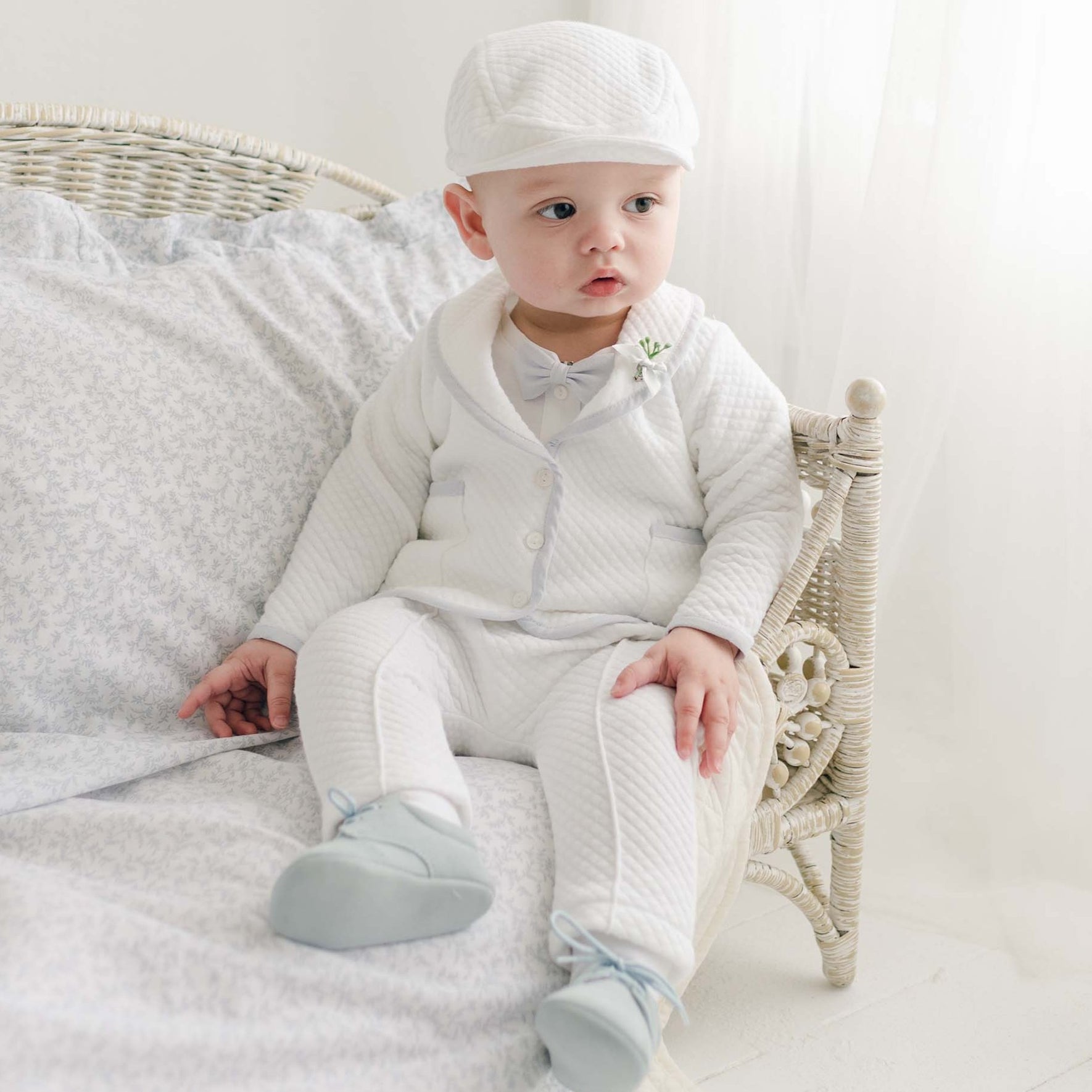 A baby boy dressed in the Harrison Christening Pants Suit is sitting on a light-colored wicker chair with cushioned bedding.