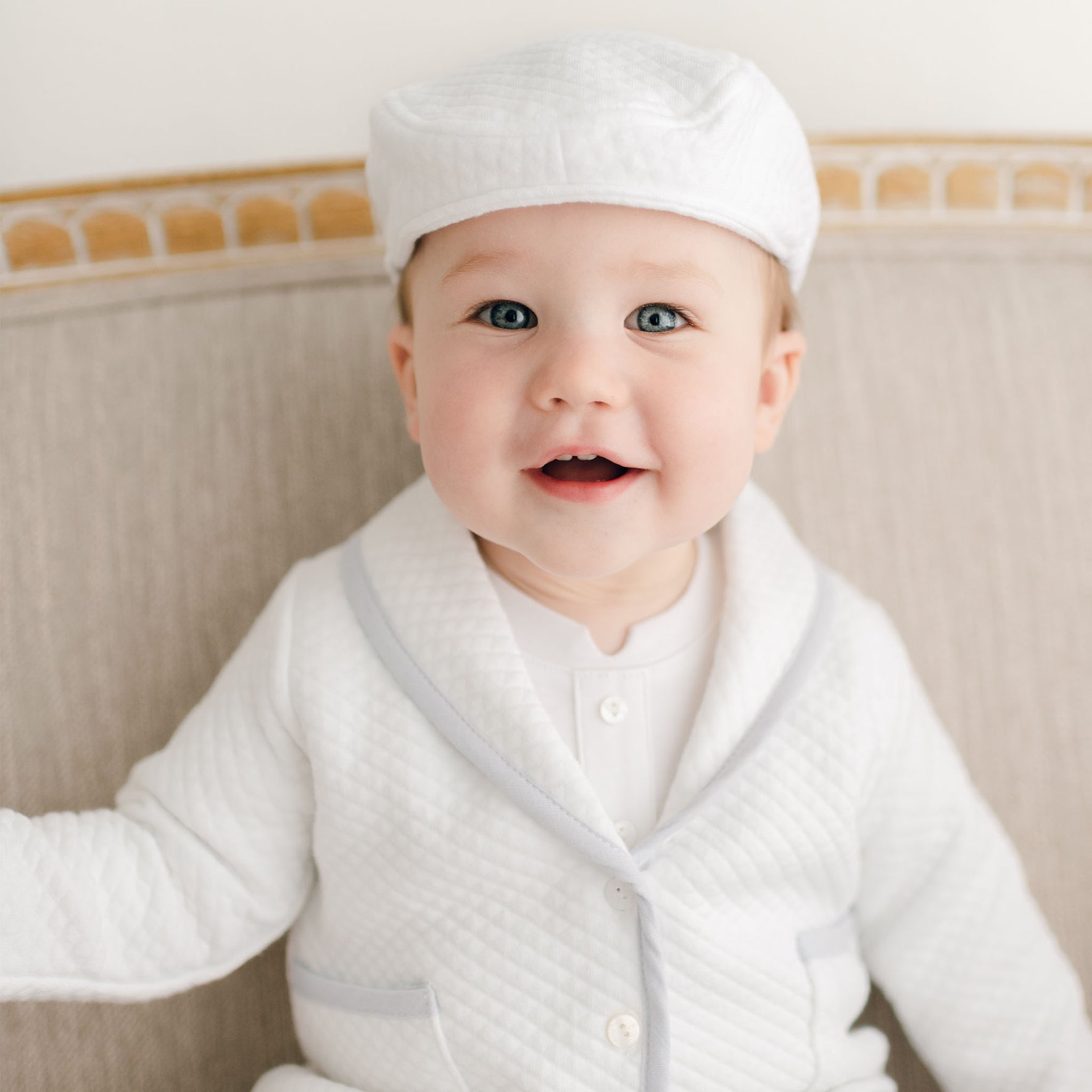Smiling baby boy in the Harrison Christening Pants Suit, styled with a quilted white jacket, light blue trim, and matching cap, seated on a light beige sofa.