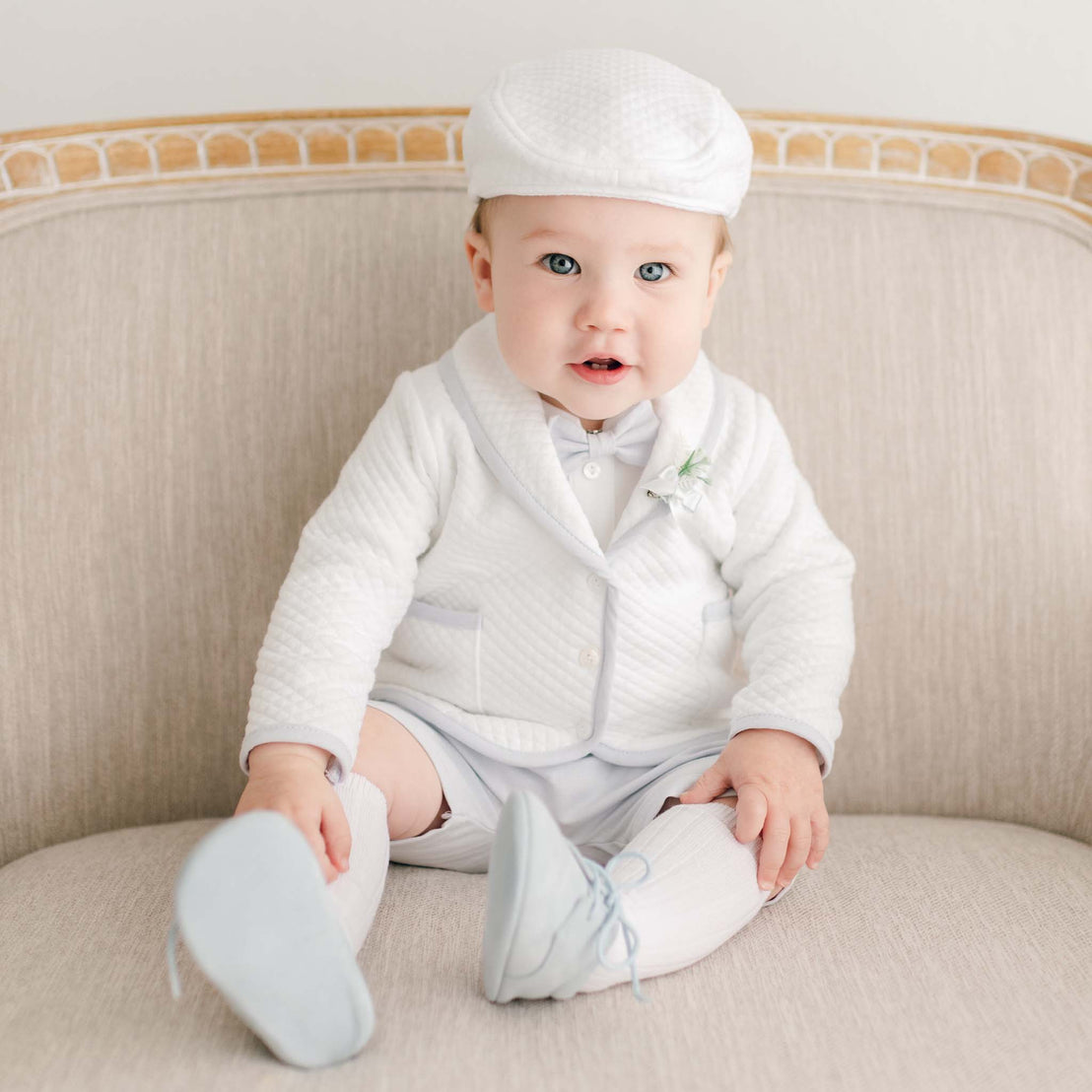 Baby boy sitting on a chair wearing the Harrison 3-Piece Shorts Suit made with plush white quilted cotton jacket, hat, and light blue shorts