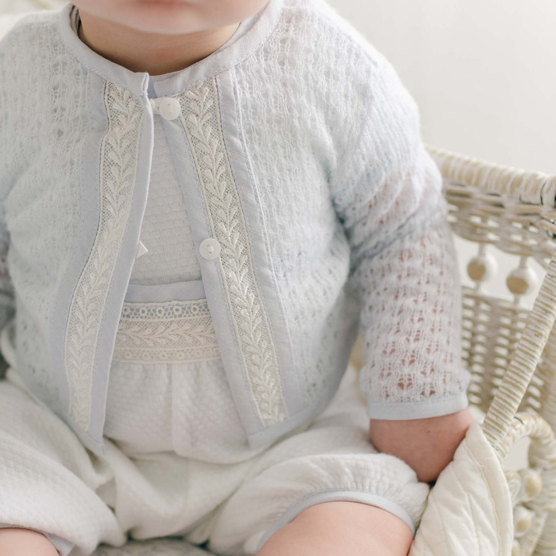 A close-up photo of a baby sitting in a woven basket. The baby is wearing a light-colored, lace-patterned Harrison Blue Knit Christening Sweater over a textured romper. The image focuses on the baby boy's comfort, emphasizing his torso with the face partially out of the frame.