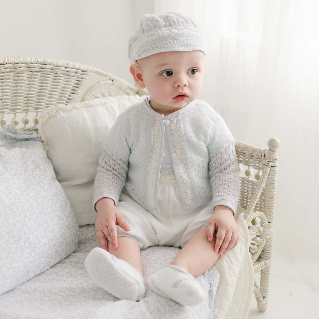 A baby dressed in a handmade in the USA blue knitted outfit and matching hat sits on a wicker chair. The baby, wearing his Harrison Blue Knit Christening Sweater, looks off to the side with a neutral expression. The softly lit background with sheer white curtains creates a serene setting.