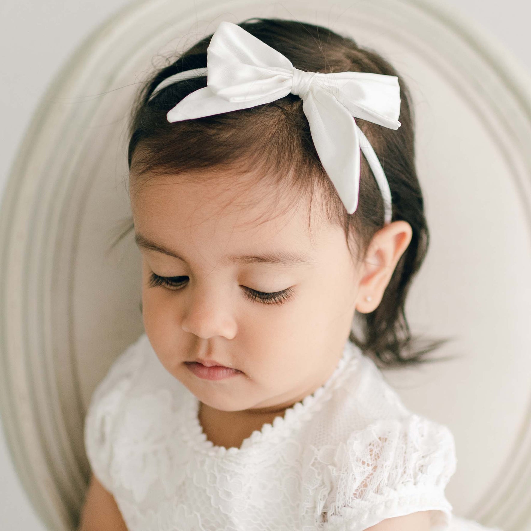 A toddler with dark hair is wearing the Victoria Silk Bow Headband in ivory. The child is looking down with a neutral expression. The background is light-colored, and the child is seated on a round-backed chair.