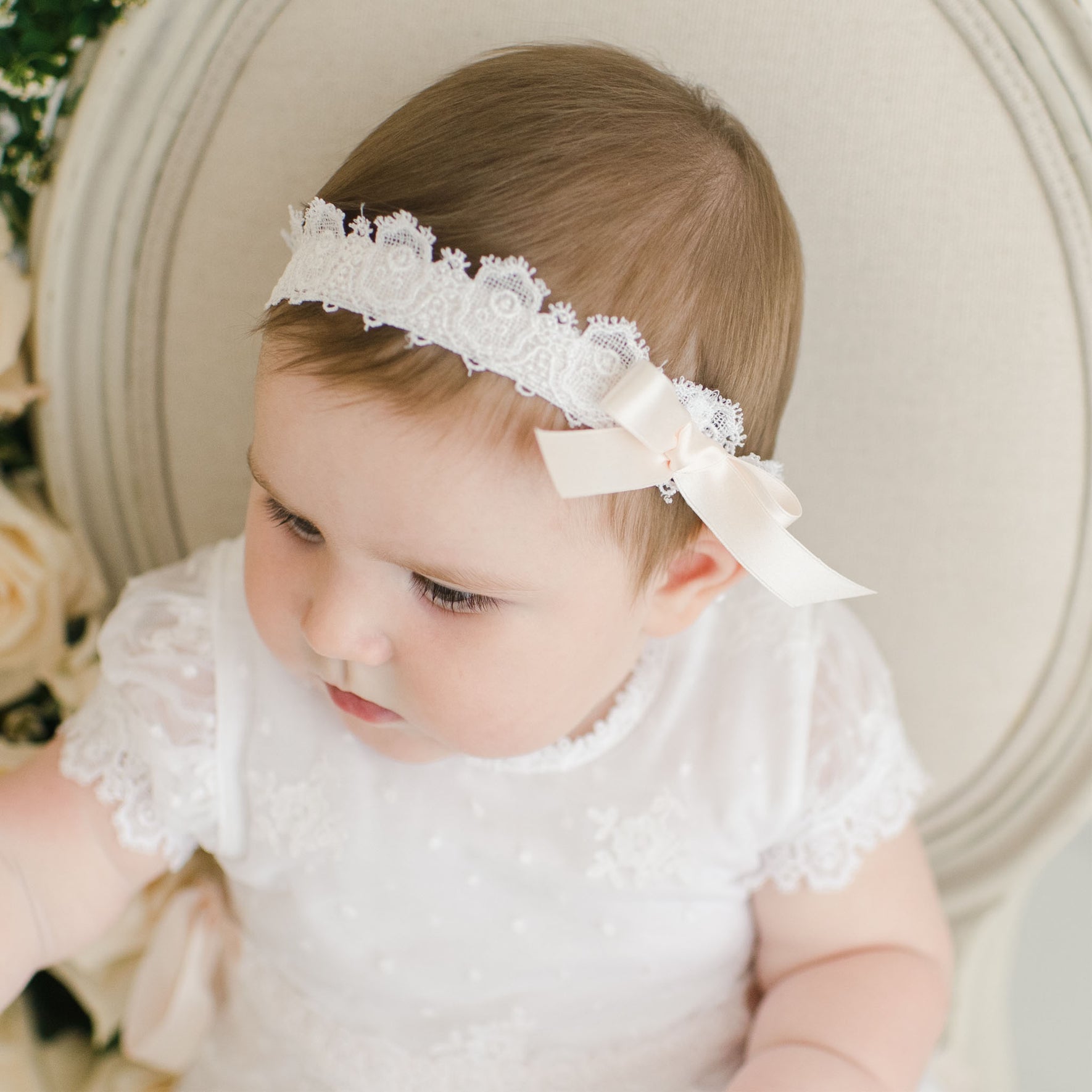 A baby wearing a white Melissa Headband with a pink silk ribbon bow sits on a light-colored chair. The baby is dressed in a white lace dress and gazes off to the side. Roses are partially visible in the background.