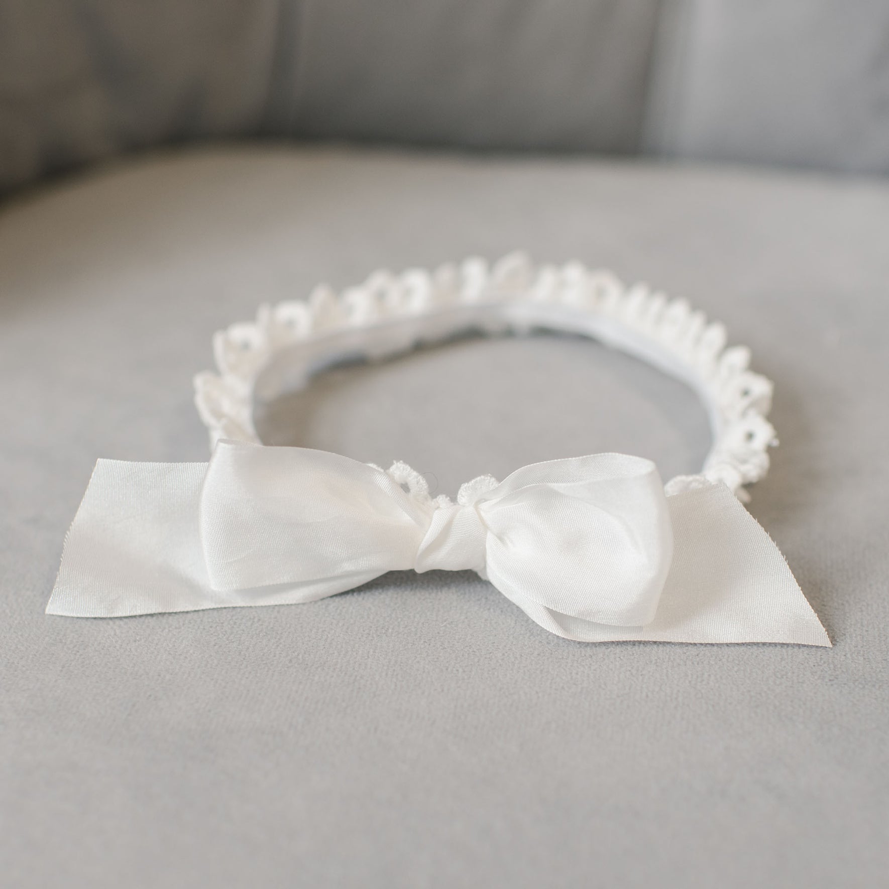 The Grace Christening Headband lies on a light gray surface, featuring a large satin bow on the front and adorned with small decorative loops around its perimeter.