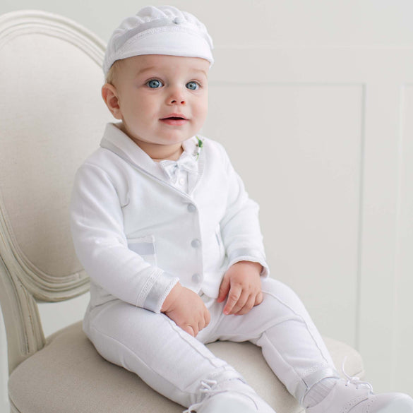 Baby in all white outfit sitting on a chair with a plain background.