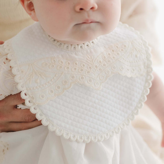 Close-up of a baby wearing the Ingrid Bib, which features intricate handmade embroidery and a vintage touch. The baby is dressed in white, with an adult's hand gently resting on their shoulder. The background is softly blurred and light-toned.